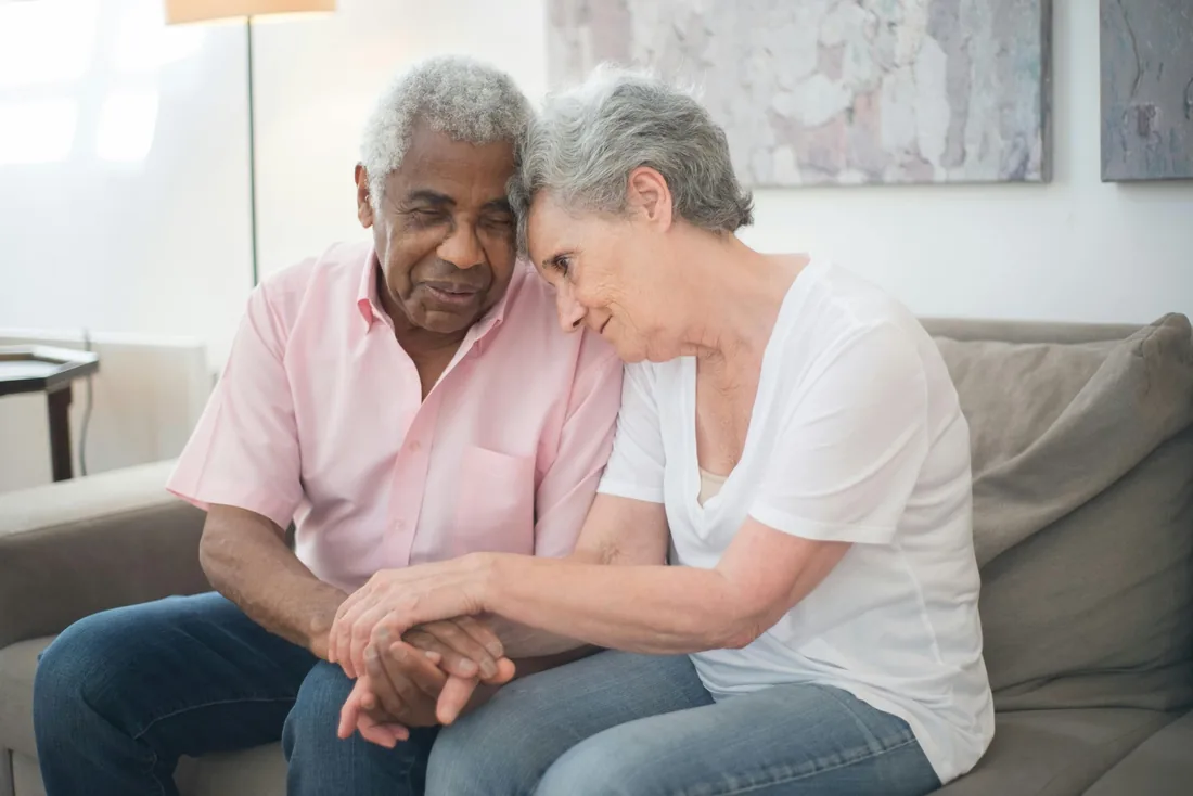 An elderly couple sitting close together on a sofa, holding hands, heads gently touching
